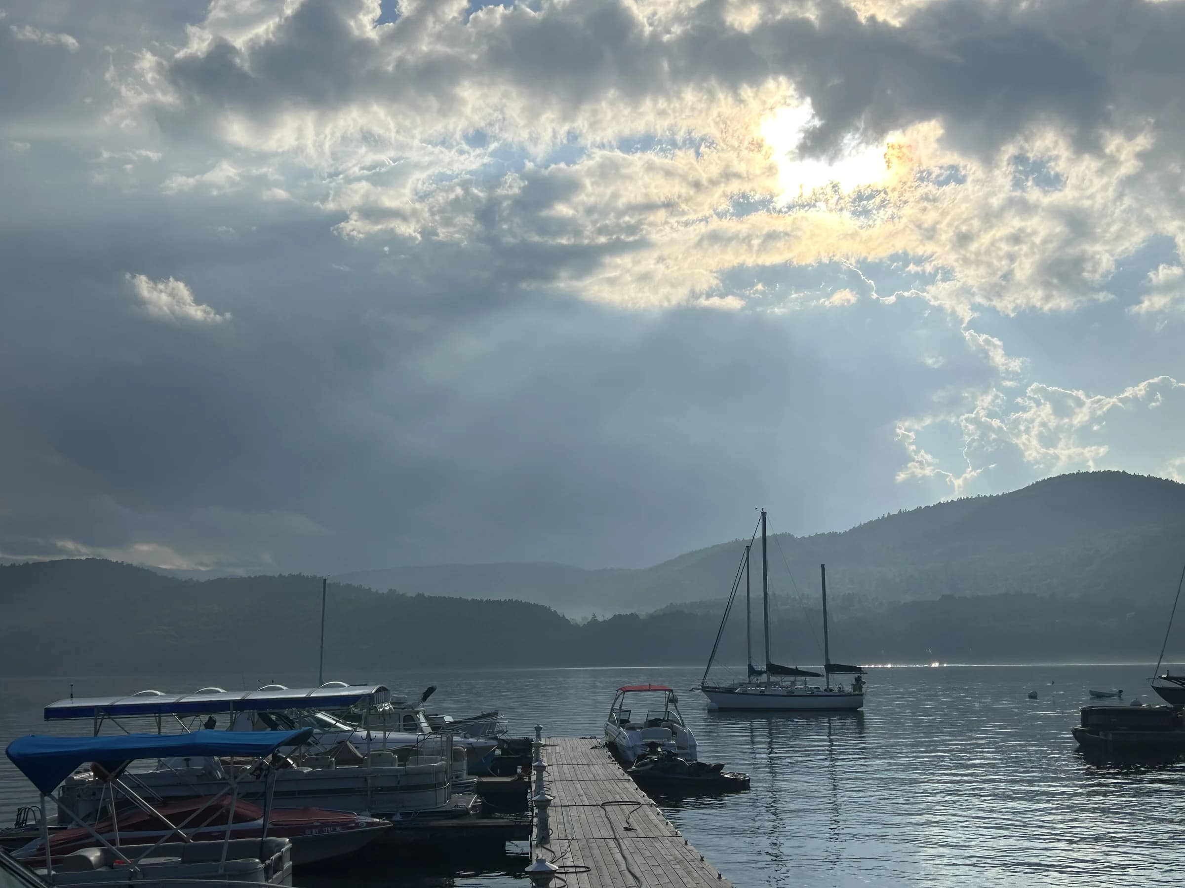 Boats moored at a marina beneath sunlit clouds and mountain ridges